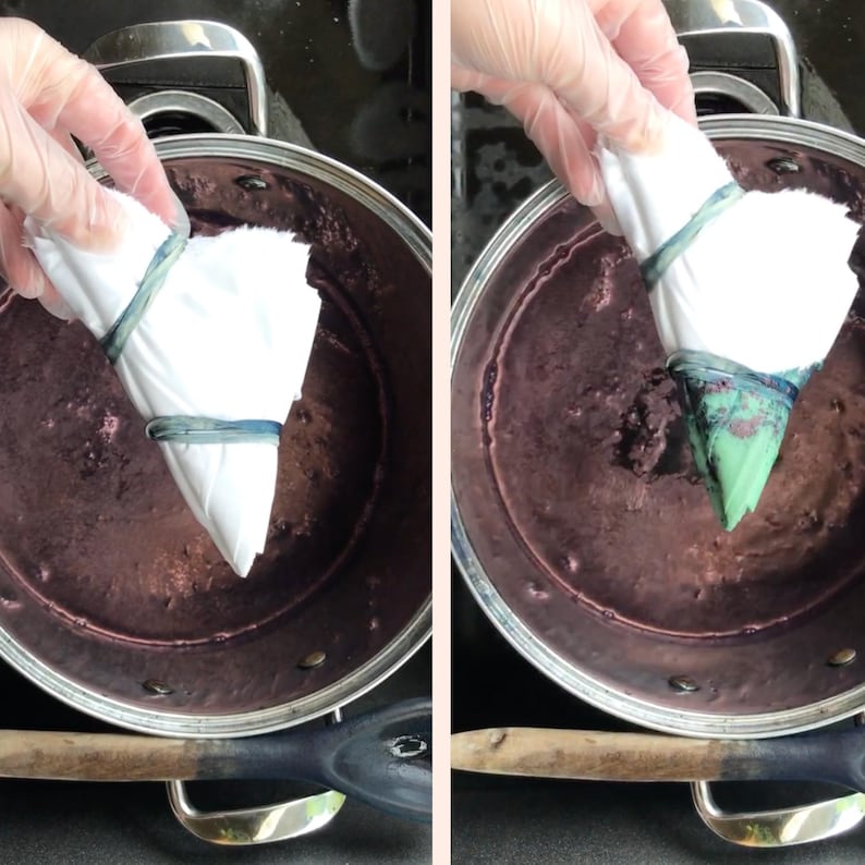 May include: A close-up of a tie-dye process. A white fabric bundle, secured with a blue rubber band, is submerged in a pot of dark purple dye. The fabric is being held by a gloved hand. The dye is a deep, rich color.