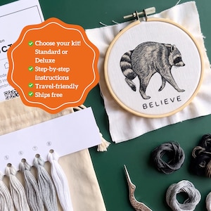 May include: An embroidery kit featuring a wooden hoop with a white fabric panel displaying a black ink illustration of a raccoon and the word BELIEVE. Includes a thread organizer with numbered white and gray embroidery floss and a metal crane scissor.