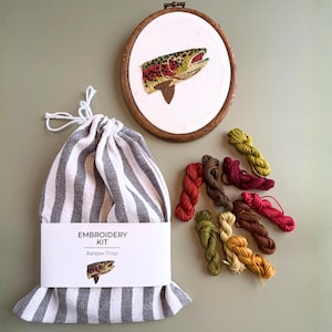 May include: An embroidery kit featuring a wooden hoop with a rainbow trout design on white fabric, a grey and white striped drawstring bag labelled EMBROIDERY KIT Rainbow Trout, and several small skeins of colourful embroidery thread in various hues.