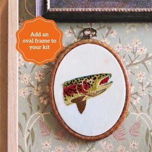 May include: An oval wooden embroidery hoop containing white fabric with a hand-stitched trout fish design. The fish features green, yellow, and red thread details. An orange graphic overlay with the text Add an oval frame to your kit is present.