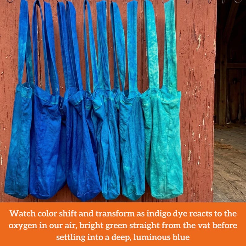 Op de afbeelding: Een rij blauwe draagtassen met lange banden hangt tegen een verweerde rode houten muur. De tassen vari&euml;ren in blauwtinten, van helder turkoois tot diep indigo. De tekst op de afbeelding luidt: "Watch color shift and transform..."