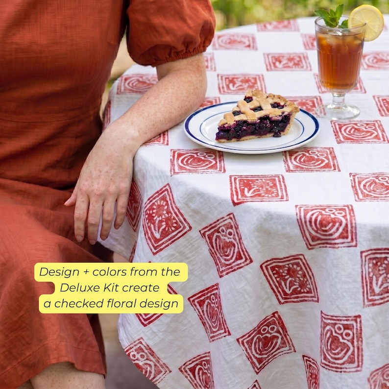 May include: A white tablecloth with a red floral pattern covers a round table. A slice of pie sits on a white plate with a blue rim. A glass of iced tea with a lemon slice and mint sprig is also on the table. The text on the image says "Design + colors from the Deluxe Kit create a checked floral design."
