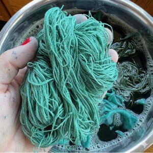Op de afbeelding: Een hand houdt een grote bundel turquoise garen vast, vers geverfd. Het garen heeft een levendige turquoise kleur, met een zacht, getextureerd uiterlijk. Het garen zit in een roestvrijstalen kom met donker turquoise kleurstof en bellen.