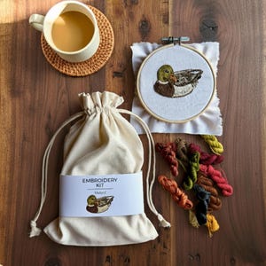 Puede incluir: Un kit de bordado con un dise&ntilde;o de pato real. El kit incluye un bastidor con el pato bordado, una bolsa beige con cord&oacute;n y hilo de bordar colorido. Una taza de caf&eacute; est&aacute; sobre un posavasos tejido.