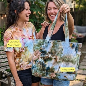 May include: Two women smiling, one wearing a Quartz Mountain tie-dye t-shirt, the other holding a Waynoka tie-dye tote bag. The tote bag has two exterior pockets and matching handles. The tie-dye pattern includes shades of blue, green, and pink.