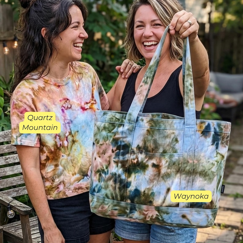 May include: Two women smiling, one wearing a Quartz Mountain tie-dye t-shirt, the other holding a Waynoka tie-dye tote bag. The tote bag has two exterior pockets and matching handles. The tie-dye pattern includes shades of blue, green, and pink.