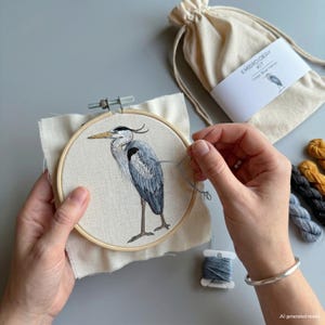 Könnte beinhalten: Ein Stickset mit einem runden Holzrahmen und einem cremefarbenen Stoffeinsatz, der einen gestickten Graureiher zeigt. Das Set enthält einen beigen Kordelzugbeutel mit der Aufschrift EMBROIDERY KIT Great Blue Heron sowie verschiedene Stickgarne.