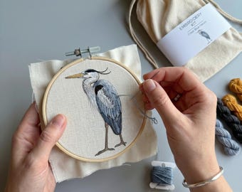 May include: An embroidery kit featuring a round wooden hoop with a cream fabric panel displaying a stitched grey heron. The kit includes a beige drawstring storage bag labeled EMBROIDERY KIT Great Blue Heron and assorted embroidery threads.