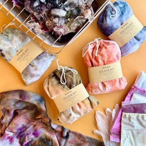 May include: An overhead shot of several ice dye kits in various colors, including blue, pink, and brown. Each kit is bundled and labeled with a brown paper band that reads "ICE DYE KIT". A metal tray filled with ice sits at the top.