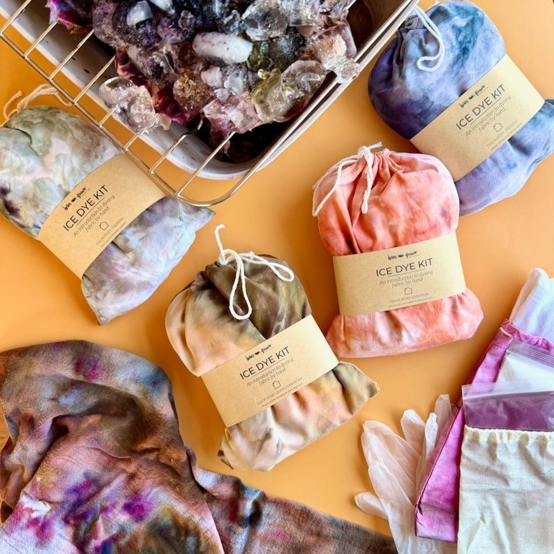May include: An overhead shot of several ice dye kits in various colors, including blue, pink, and brown. Each kit is bundled and labeled with a brown paper band that reads "ICE DYE KIT". A metal tray filled with ice sits at the top.