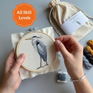 May include: An embroidery kit featuring a round wooden hoop with a grey heron design stitched on white fabric, a needle, a beige drawstring storage bag labeled EMBROIDERY KIT Great Blue Heron, and several small skeins of colored embroidery thread.