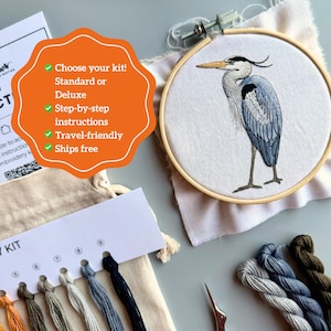 May include: An embroidery kit featuring a heron design. The heron is depicted in shades of blue, gray, and black, stitched on white fabric within a wooden hoop. Additional elements include embroidery floss in various colors and a small pair of scissors.