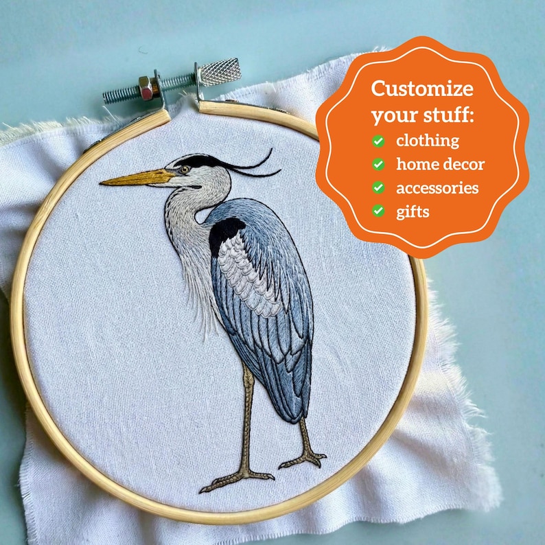 Pu&ograve; includere: Un telaio da ricamo in legno contenente tessuto bianco con un airone grigio ricamato a filo. Una sovrapposizione grafica arancione include il testo: Customize your stuff: clothing, home decor, accessories, gifts, ciascuno con una spunta verde.