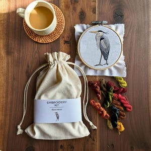 Pu&ograve; includere: Un kit da ricamo con un telaio in tessuto bianco con un'illustrazione di un airone azzurro, una sacca beige con coulisse etichettata EMBROIDERY KIT Blue Heron e matassine di filo da ricamo colorate accanto a una tazza su un sottobicchiere.
