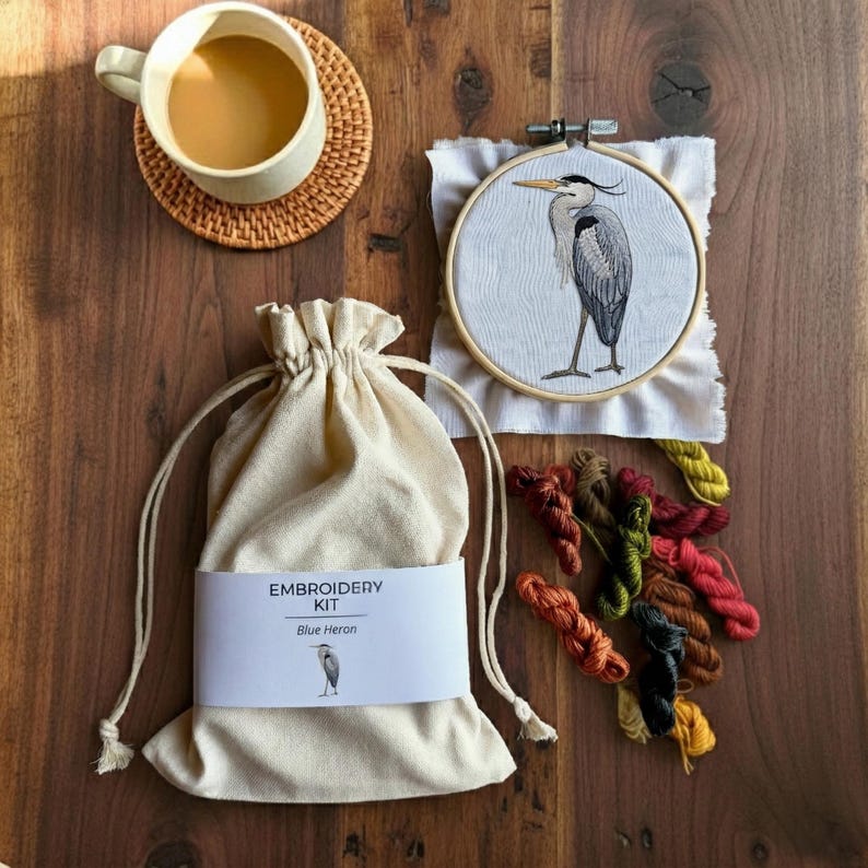 Pu&ograve; includere: Un kit da ricamo con un telaio in tessuto bianco con un'illustrazione di un airone azzurro, una sacca beige con coulisse etichettata EMBROIDERY KIT Blue Heron e matassine di filo da ricamo colorate accanto a una tazza su un sottobicchiere.