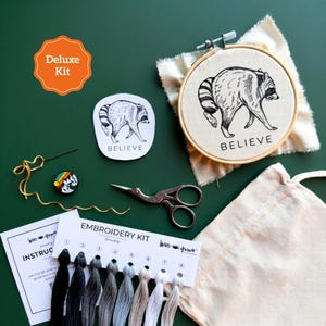 May include: An embroidery kit featuring a wooden hoop with a raccoon illustration and the word BELIEVE, a matching sticker, embroidery floss in eight shades from black to white, a needle, scissors, a small round pin, and an instruction card.