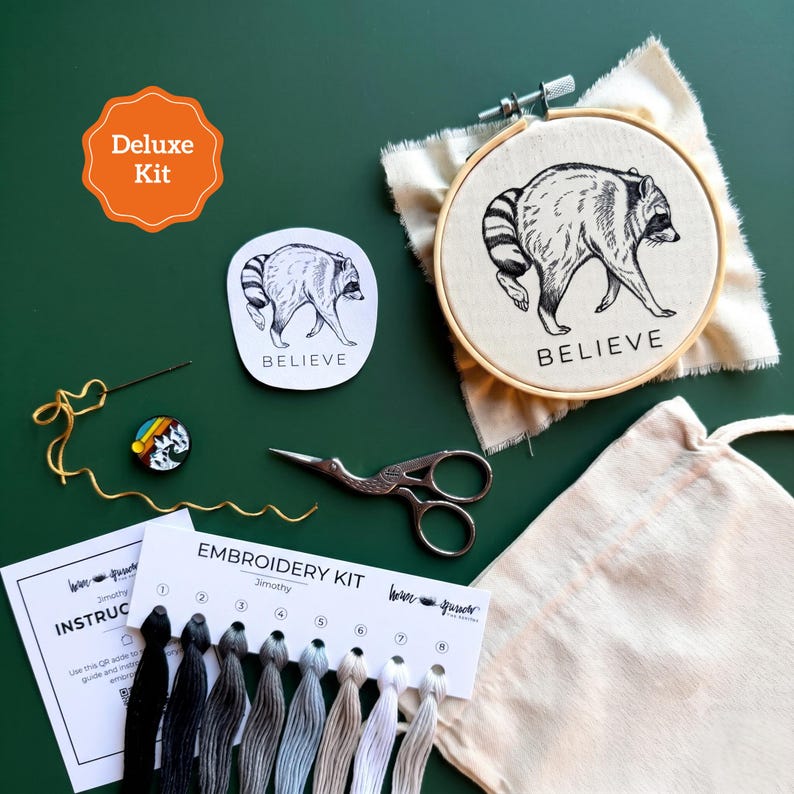 May include: An embroidery kit featuring a wooden hoop with a raccoon illustration and the word BELIEVE, a matching sticker, embroidery floss in eight shades from black to white, a needle, scissors, a small round pin, and an instruction card.