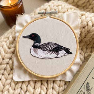 May include: A circular wooden embroidery hoop containing a piece of off-white fabric with a detailed thread-stitched design of a common loon. The bird features a black head with a red eye, a white neck patch, and black and white patterned wings.