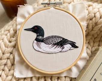 May include: A circular wooden embroidery hoop containing a piece of off-white fabric with a detailed thread-stitched design of a common loon. The bird features a black head with a red eye, a white neck patch, and black and white patterned wings.