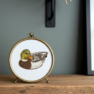 Puede incluir: Bordado enmarcado de forma redonda y color dorado de un pato real. El pato est&aacute; representado en tonos verdes, marrones y grises, con un pico naranja. El marco tiene un peque&ntilde;o soporte y se exhibe sobre una superficie de madera.