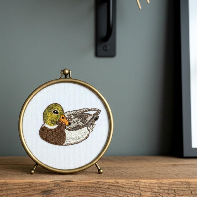 Puede incluir: Bordado enmarcado de forma redonda y color dorado de un pato real. El pato est&aacute; representado en tonos verdes, marrones y grises, con un pico naranja. El marco tiene un peque&ntilde;o soporte y se exhibe sobre una superficie de madera.