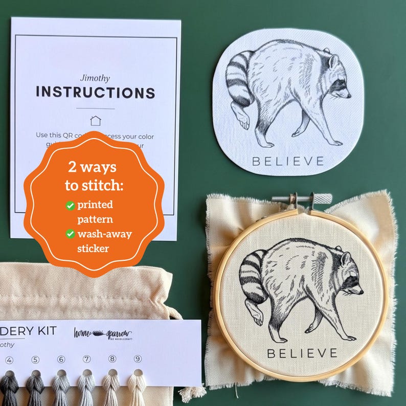 May include: An embroidery kit featuring a round wooden hoop with a raccoon illustration and the word BELIEVE, a matching circular sticker, an instruction card with QR code, a thread organizer with numbered slots, and a small beige fabric pouch.