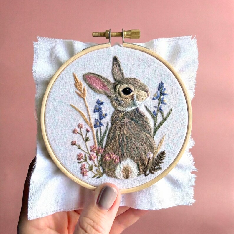 May include: A circular wooden embroidery hoop containing white fabric with a hand-stitched design of a brown rabbit surrounded by small pink flowers, blue wildflowers, and tall golden wheat stalks. The rabbit has a white tail and pink inner ears.