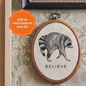 May include: An oval wooden embroidery hoop containing a light-colored fabric panel with a black and white stitched illustration of a raccoon walking. The word BELIEVE is embroidered in black capital letters below the raccoon figure.