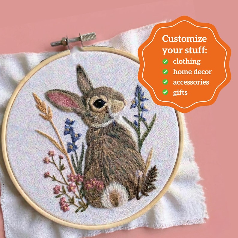 May include: A round wooden embroidery hoop containing white fabric with a detailed thread-stitched brown rabbit surrounded by blue and pink wildflowers. An orange scalloped graphic includes the text: Customize your stuff: clothing, home decor, accessories, gifts.