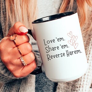 Könnte beinhalten: Eine weiße Keramiktasse mit schwarzem Rand und Henkel. Die Tasse hat ein rosafarbenes Herzdesign und den Text "Love 'em. Share 'em. Reverse Harem."