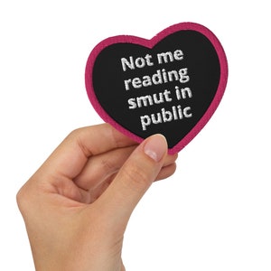 以下が含まれることがあります： 「Not me reading smut in public.」と刺繍された、黒地に白の刺繍が施されたピンクのハート型のワッペンです。