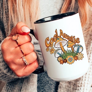 May include: White ceramic mug with a black rim and handle. The mug has a graphic of mushrooms and flowers with the text "Cool Auntie" in yellow and black.