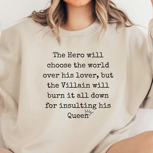May include: A light beige sweatshirt with the text "The Hero will choose the world over his lover, but the Villain will burn it all down for insulting his Queen" printed in black.