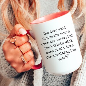 May include: White ceramic mug with a pink rim and handle. The mug has black text that reads "The Hero will choose the world over his lover, but the Villain will burn it all down for insulting his Queen".