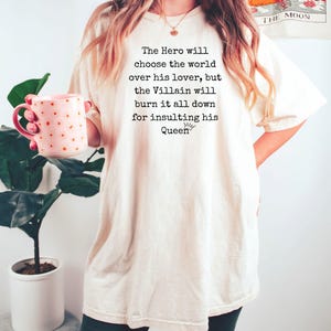May include: Cream-colored t-shirt with the text "The Hero will choose the world over his lover, but the Villain will burn it all down for insulting his Queen." A pink mug with gold polka dots is held in the left hand.