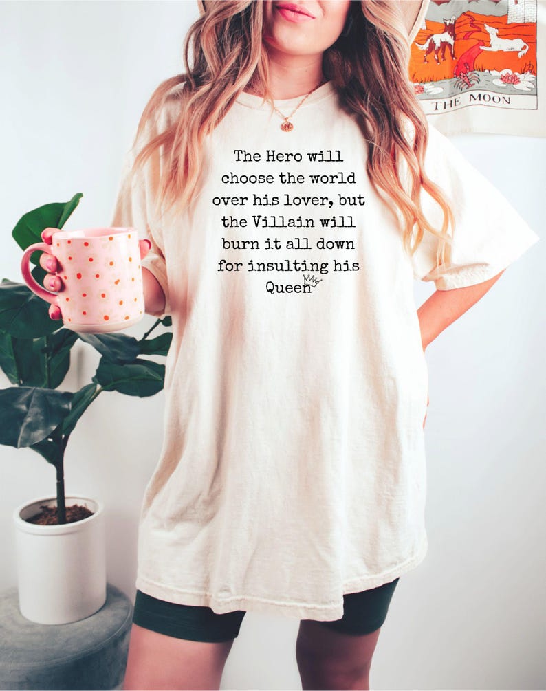 May include: Cream-colored t-shirt with the text "The Hero will choose the world over his lover, but the Villain will burn it all down for insulting his Queen." A pink mug with gold polka dots is held in the left hand.