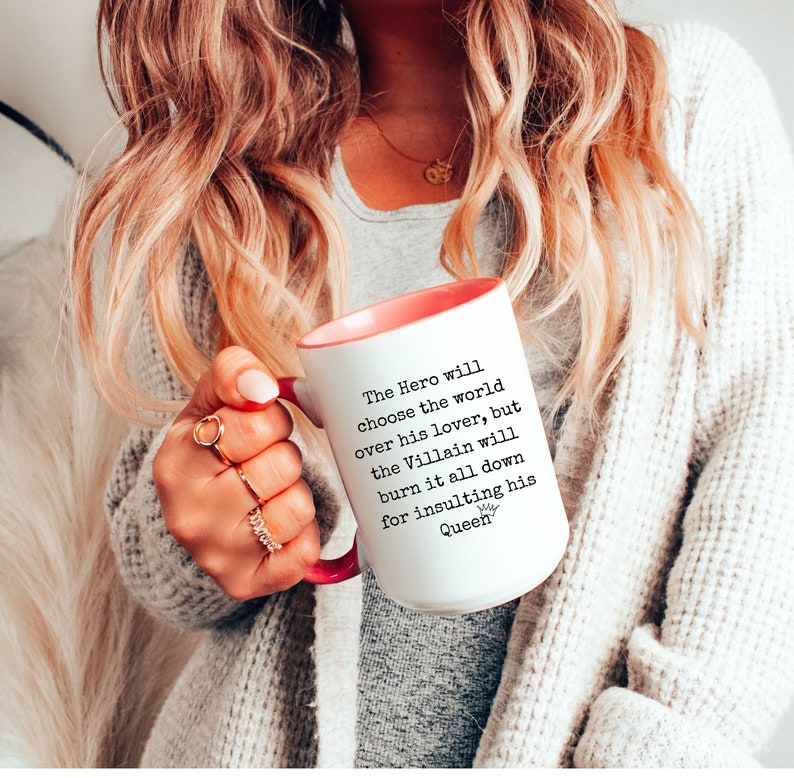 May include: White ceramic mug with a pink rim and handle. The mug has black text that reads "The Hero will choose the world over his lover, but the Villain will burn it all down for insulting his Queen".