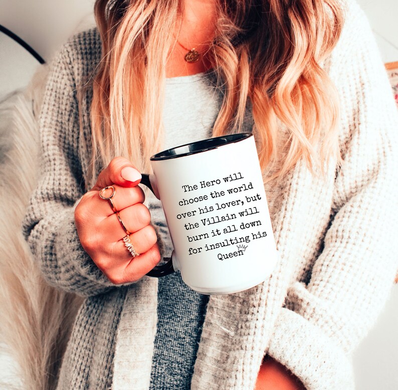 May include: White ceramic mug with a black rim and handle. The mug has black text that reads "The Hero will choose the world over his lover, but the Villain will burn it all down for insulting his Queen".