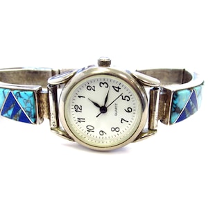 Op de afbeelding: Zilverkleurig quartz horloge met een witte wijzerplaat en zwarte cijfers. De horlogeband heeft turkoois en donkerblauwe steen inlays. Een modieus accessoire.