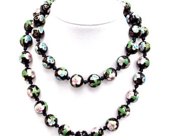 May include: Two strands of a black beaded necklace with floral designs in pink, blue, and green. The beads are round and appear to be made of enamel. The necklace is displayed on a white mannequin.