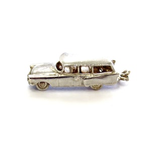 May include: A silver-colored vintage car charm with a detailed design. The charm features a classic car shape with visible windows and wheels. The car has a loop for attaching to a bracelet or necklace. The charm is made of a shiny metal.