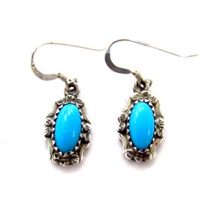 May include: A pair of silver drop earrings featuring oval, bright turquoise stones. The stones are set in ornate, silver frames with decorative edges. The earrings have a classic, elegant design.