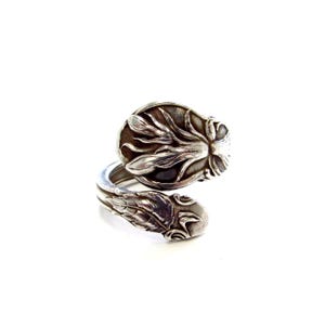 May include: A silver-toned bypass ring crafted from a repurposed spoon handle. The ring features an embossed floral pattern with intricate leaf details and a polished finish, showcasing a wrap-around design that overlaps at the center point.