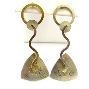 Puede incluir: Un par de pendientes de color dorado con un colgante triangular y una barra ondulada y curva. Los colgantes tienen una superficie texturizada con un diseño circular y un patrón de espiral.