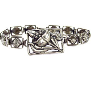 May include: A silver chain bracelet featuring a floral design. The flower is a stylised tulip with leaves and a bud. The chain is made of small, square links.