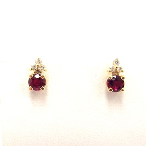 Puede incluir: Un par de pendientes de botón de oro, cada uno con una piedra preciosa redonda de color rojo intenso en la base, rematada con un grupo de tres piedras pequeñas, claras y brillantes. El engaste de oro amarillo ofrece un contraste cálido.