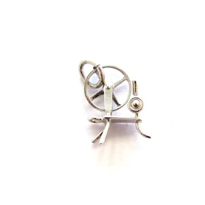 May include: A silver-colored charm in the shape of a spinning wheel. The charm has a circular wheel with spokes, a small platform, and a curved leg. The charm is likely made of metal and is on a white background.