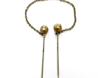 Op de afbeelding: Een paar goudkleurige haarspelden met decoratieve bolvormige toppen verbonden door een ketting.