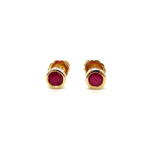 May include: A pair of stud earrings featuring round, deep pink rubies set in polished yellow gold bezels. Each earring has a smooth, circular gold frame surrounding the vibrant gemstone, creating a simple and classic design for these jewelry pieces.