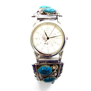 May include: Silver-toned watch with a white face featuring Roman numerals and a Native American profile. The watch band is adorned with turquoise stones and decorative silver elements. The watch has a classic design with a touch of Southwestern flair.